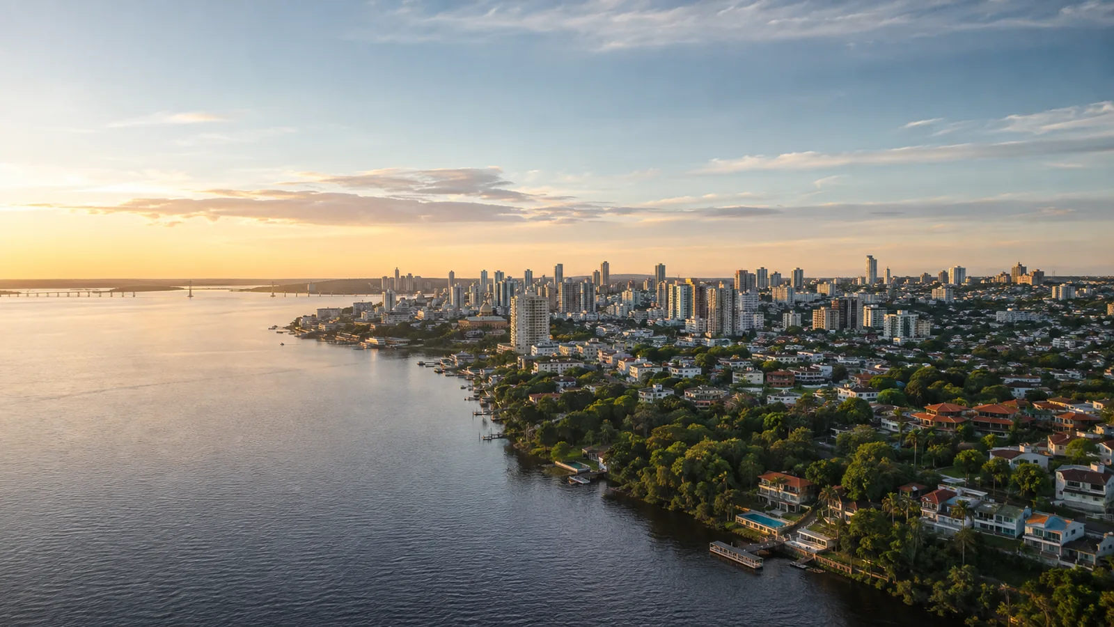 Vista aérea de uma cidade às margens de um rio no fim da tarde, com prédios, orla e área arborizada